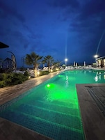 A nighttime pool area with a green light, palm trees, and lounge chairs.
