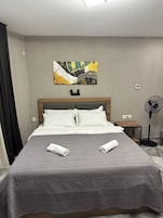 A hotel room with a bed, a bedside table, a standing fan, and a painting on the wall.