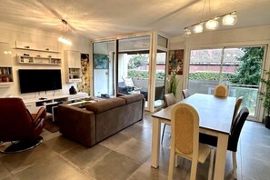 Ein modernes Wohnzimmer mit Flachbildfernseher, braunem Sofa, Esstisch mit Stühlen und einer Glastür zum Balkon.