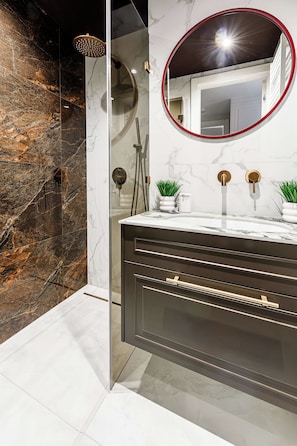 A modern bathroom with a marble wall, a round mirror, a sink with two gold faucets, and a glass-enclosed shower area.