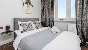 En snyggt bäddad säng med ett grått överkast, en grå sänggavel och dekorativa kuddar. Bredvid sängen står ett litet sidobord med en lampa, och ovanför bordet hänger en tavla på väggen.