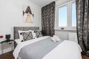 A neatly made bed with a grey blanket, a grey headboard, and decorative pillows. There is a small bedside table with a lamp and a wall art piece above it.