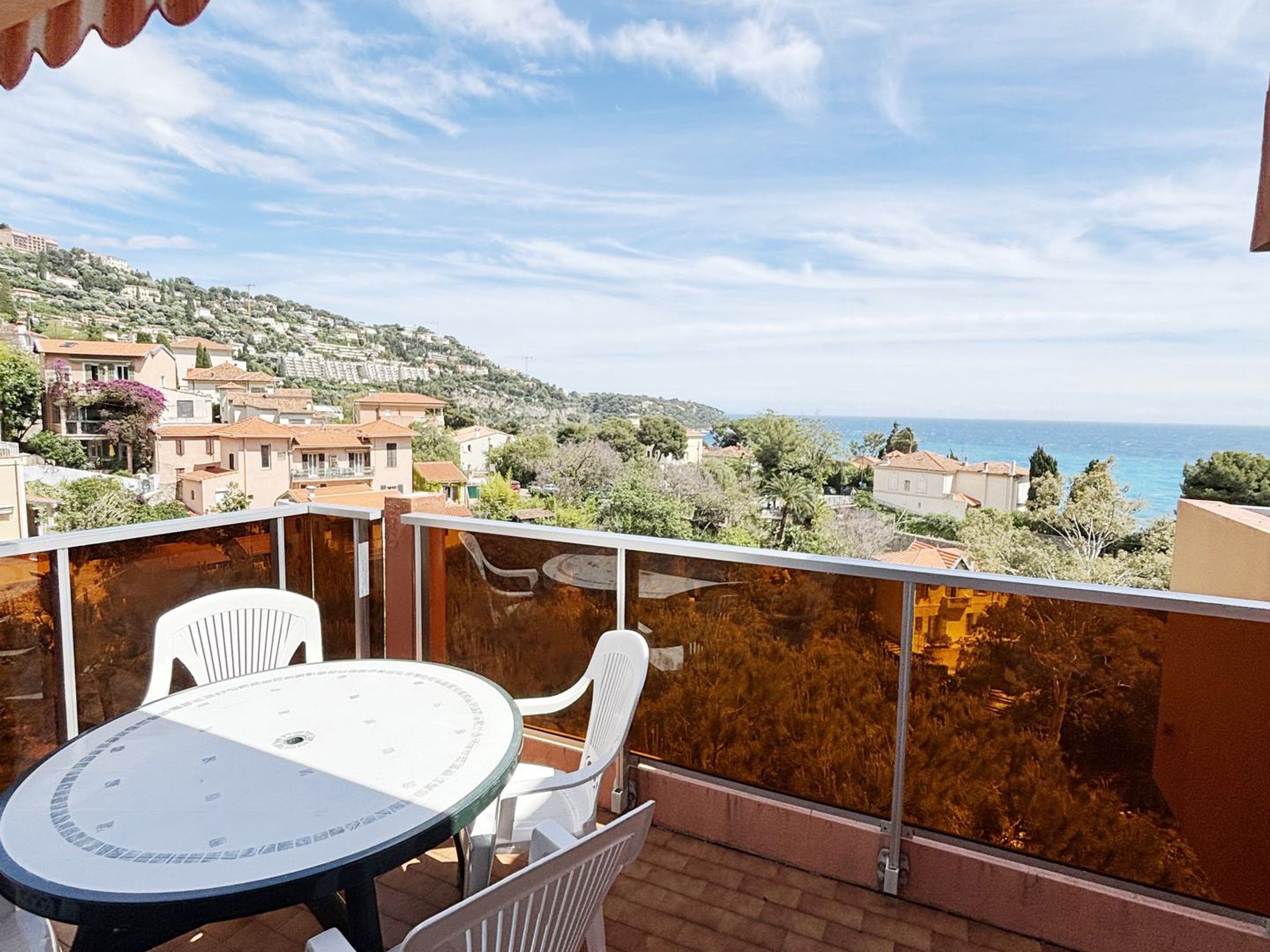 A balcony with a table and chairs, overlooking a coastal town with buildings and the sea.