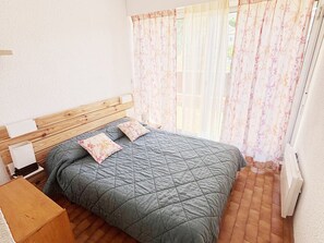 A bedroom with a wooden headboard, a bed with a quilted grey bedspread, two pillows, a bedside table with a lamp, and a window with floral curtains. - 6-person Apartment with Wi-Fi in Roquebrune-Cap-Martin (Roquebrune-Cap-Martin)
