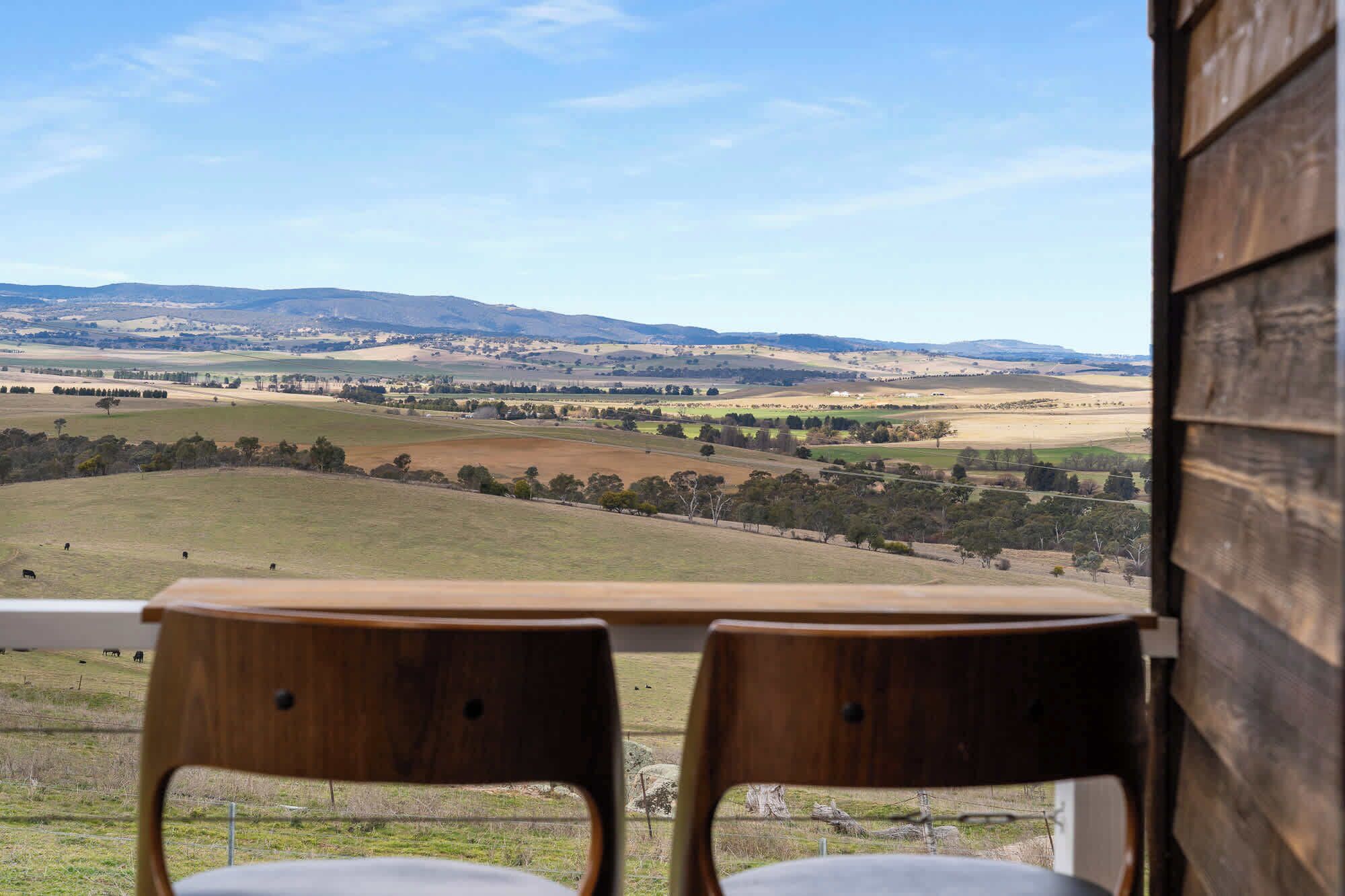 A view from a wooden-framed window overlooking a rural landscape with rolling hills and scattered trees.