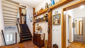 A wooden staircase leading to a second floor, a wooden cabinet with a vase of flowers, and a wall-mounted shelf with various items.