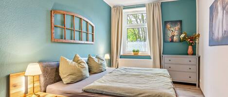 Ein Schlafzimmer mit einem hölzernen Bett, einem Nachttisch mit Lampe, einer Kommode, einem Fenster mit Vorhängen und einem Bild an der Wand.