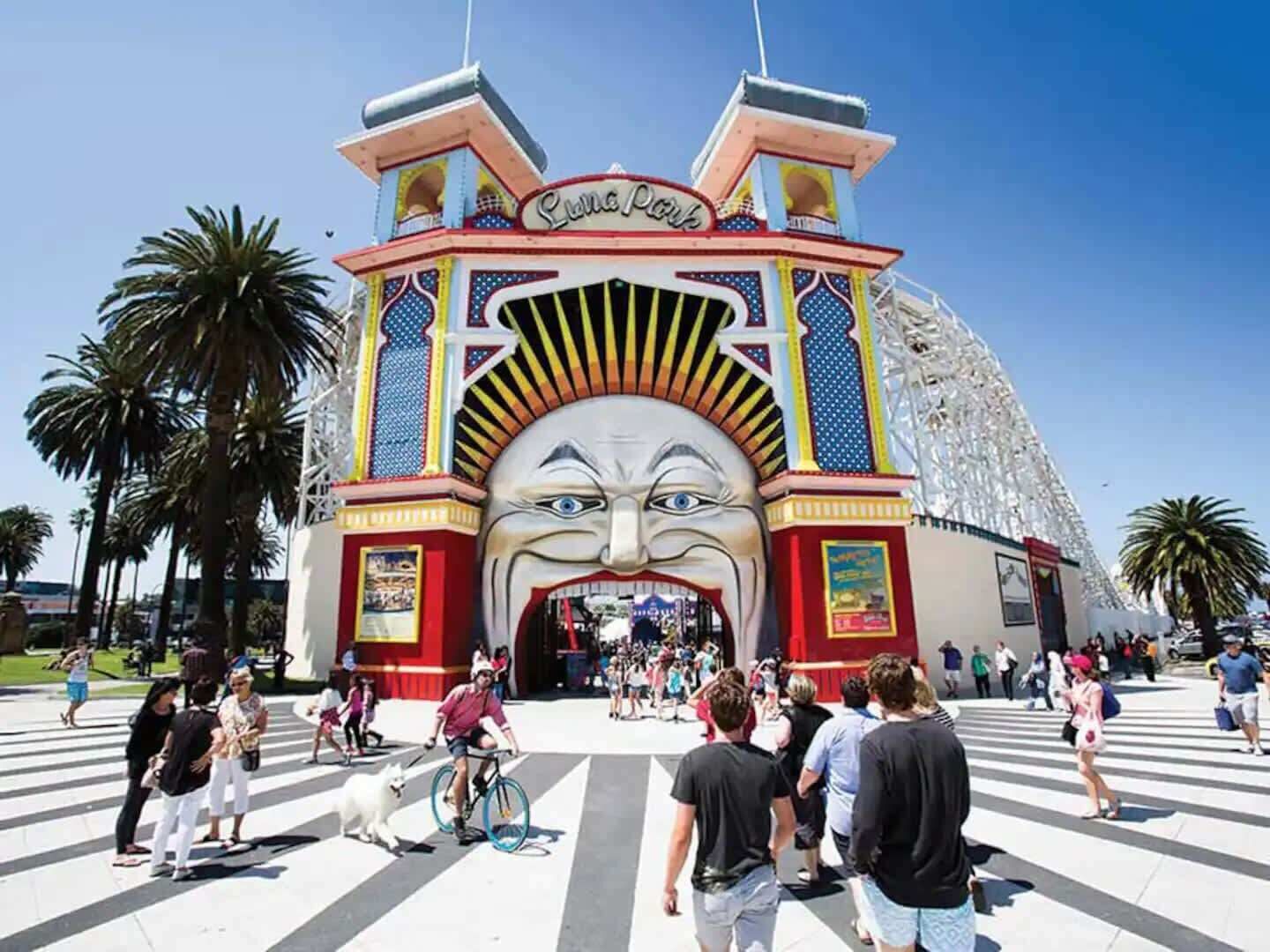 Una entrada colorida con un mural de una gran cara, palmeras y gente caminando.