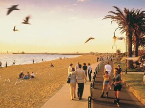 A beach scene with people walking and sitting on the sand, a pathway, and palm trees. - Luxury Stay Near Grand Prix Track (Melbourne)