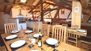 A rustic dining area with wooden tables and chairs, a kitchen in the background, and exposed wooden beams. - Chalet Bronziers - authentic alpine experience (10p, sauna) (Peisey-Nancroix)