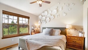 A bedroom with a bed, two bedside tables, a ceiling fan, a large window, and wall art. - 177 Wildflower - Mountain Home with New Hot Tub (Fraser)