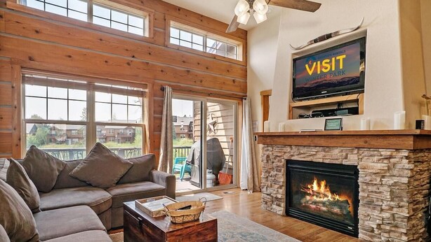 A spacious living room with a fireplace, a television mounted above it, and a large window overlooking a deck. - 177 Wildflower - Mountain Home with New Hot Tub (Fraser)