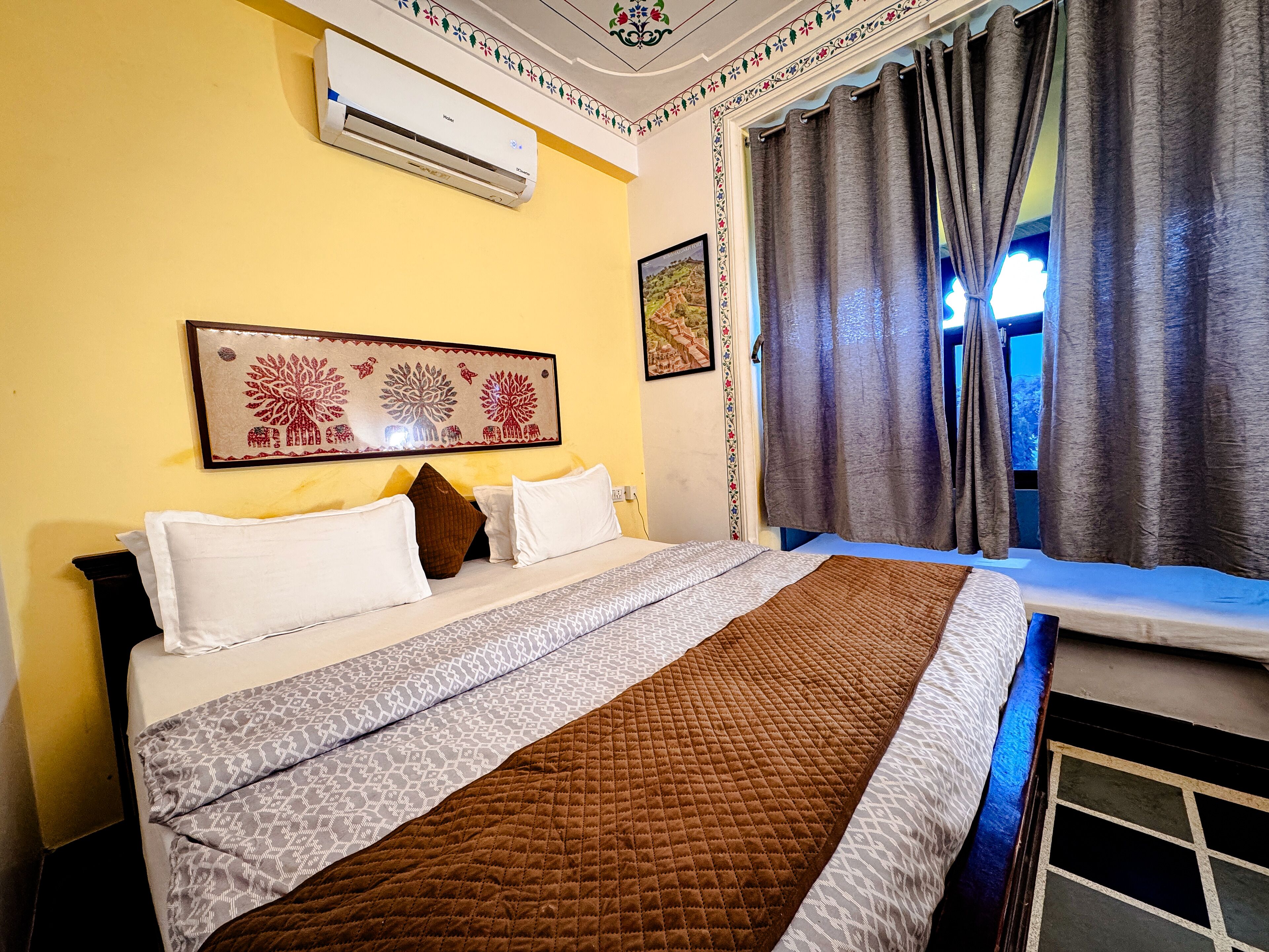 A hotel room with a bed, a window with curtains, an air conditioner, and a decorative wall piece.