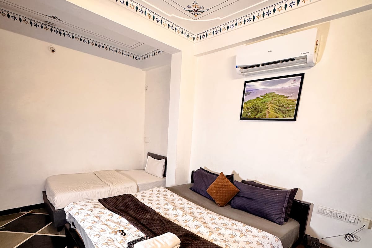A hotel room with a bed, a bedside table, a wall-mounted air conditioner, and a picture of a landscape.