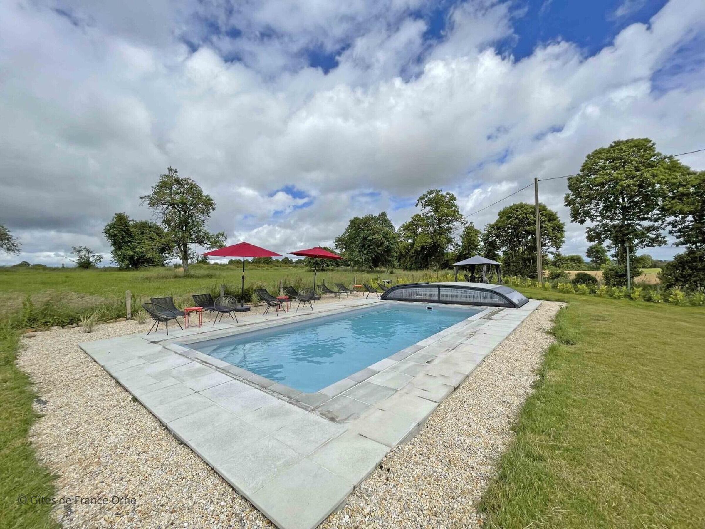 A rectangular swimming pool with a paved deck, surrounded by a gravel border. There are lounge chairs and red umbrellas in the background.