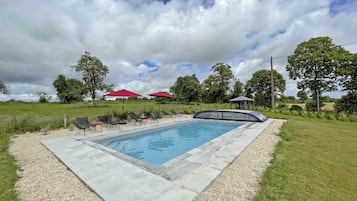 Une piscine rectangulaire entourée d’une terrasse dallée et bordée de gravier. On aperçoit, au fond, des chaises longues et des parasols rouges.