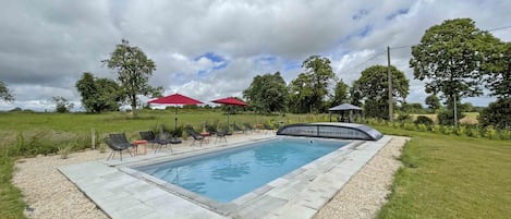 A rectangular swimming pool with a paved deck, surrounded by a gravel border. There are lounge chairs and red umbrellas in the background.