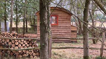 A rustic wooden cabin surrounded by trees, with a stack of logs nearby.