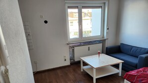A room with a wooden floor, a white table with a glass, a blue sofa, and a window with a view of buildings outside. - Furnished 1-room apartment in Bad Wörishofen (Bad Wörishofen)