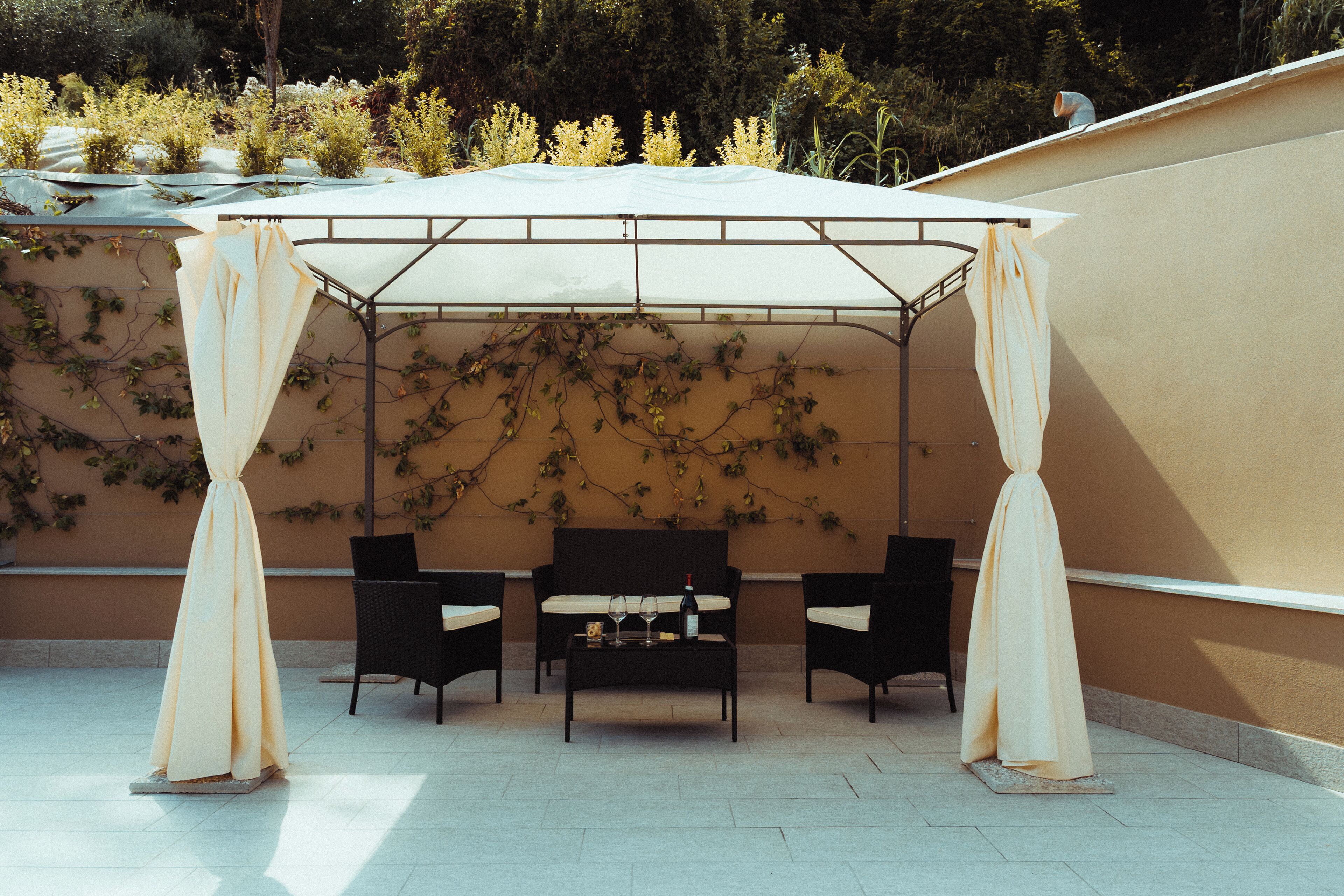 A covered outdoor seating area with two chairs and a table, surrounded by a beige wall with climbing plants.