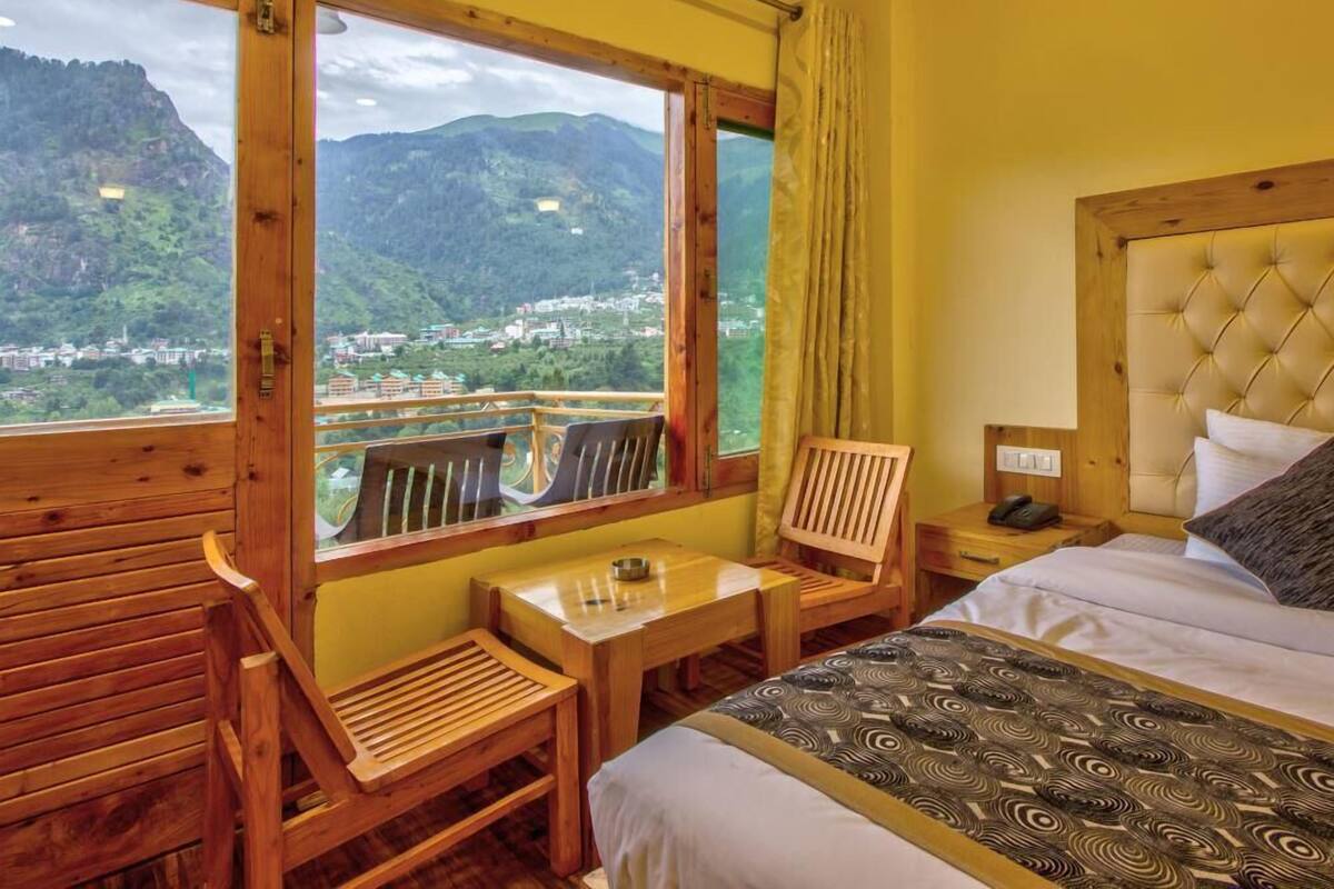 A room with a bed, a wooden table, and a chair, with a view of mountains and a town.