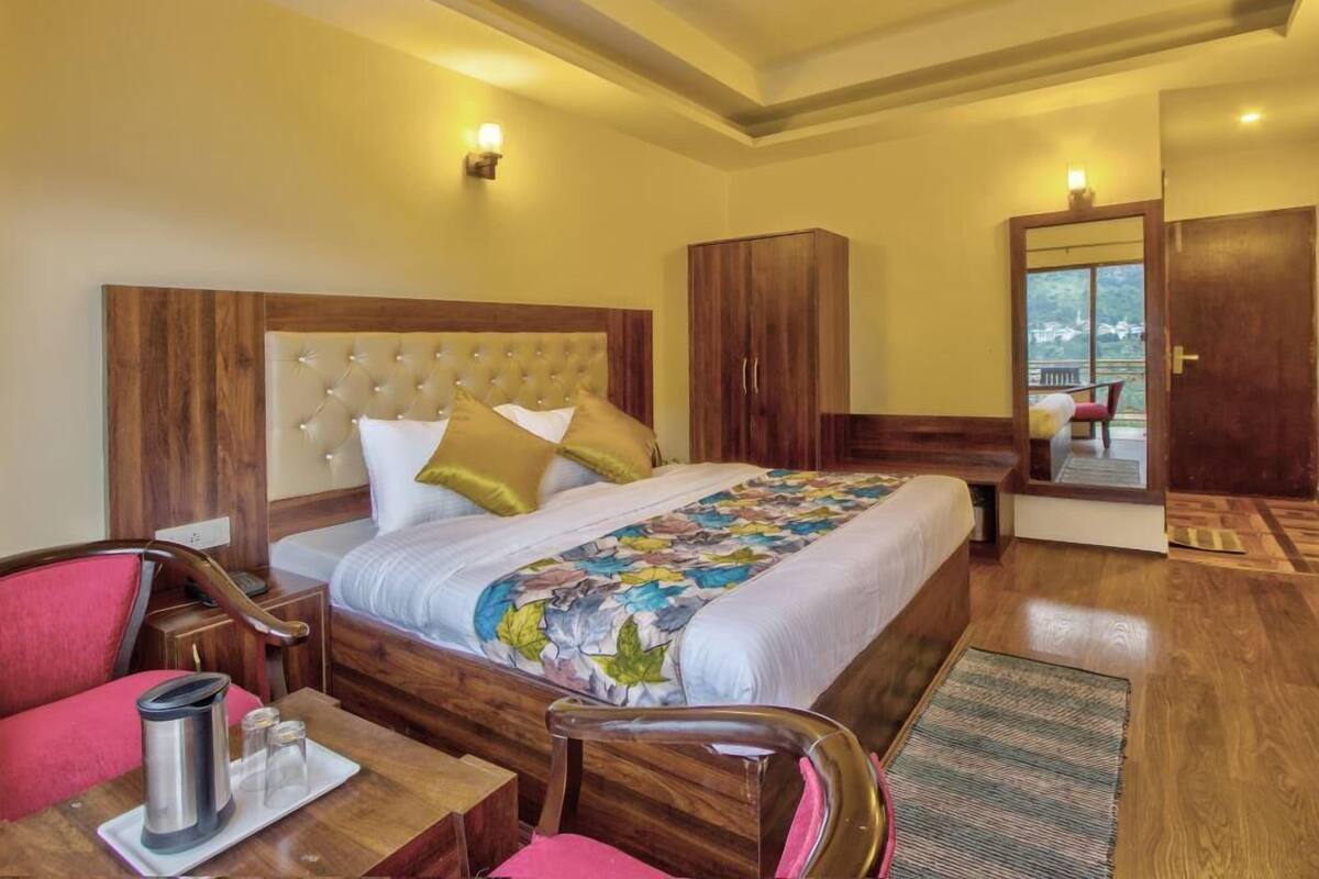 A hotel room with a large bed, wooden headboard, a wardrobe, a desk with a chair, and a small table with a kettle and glasses.