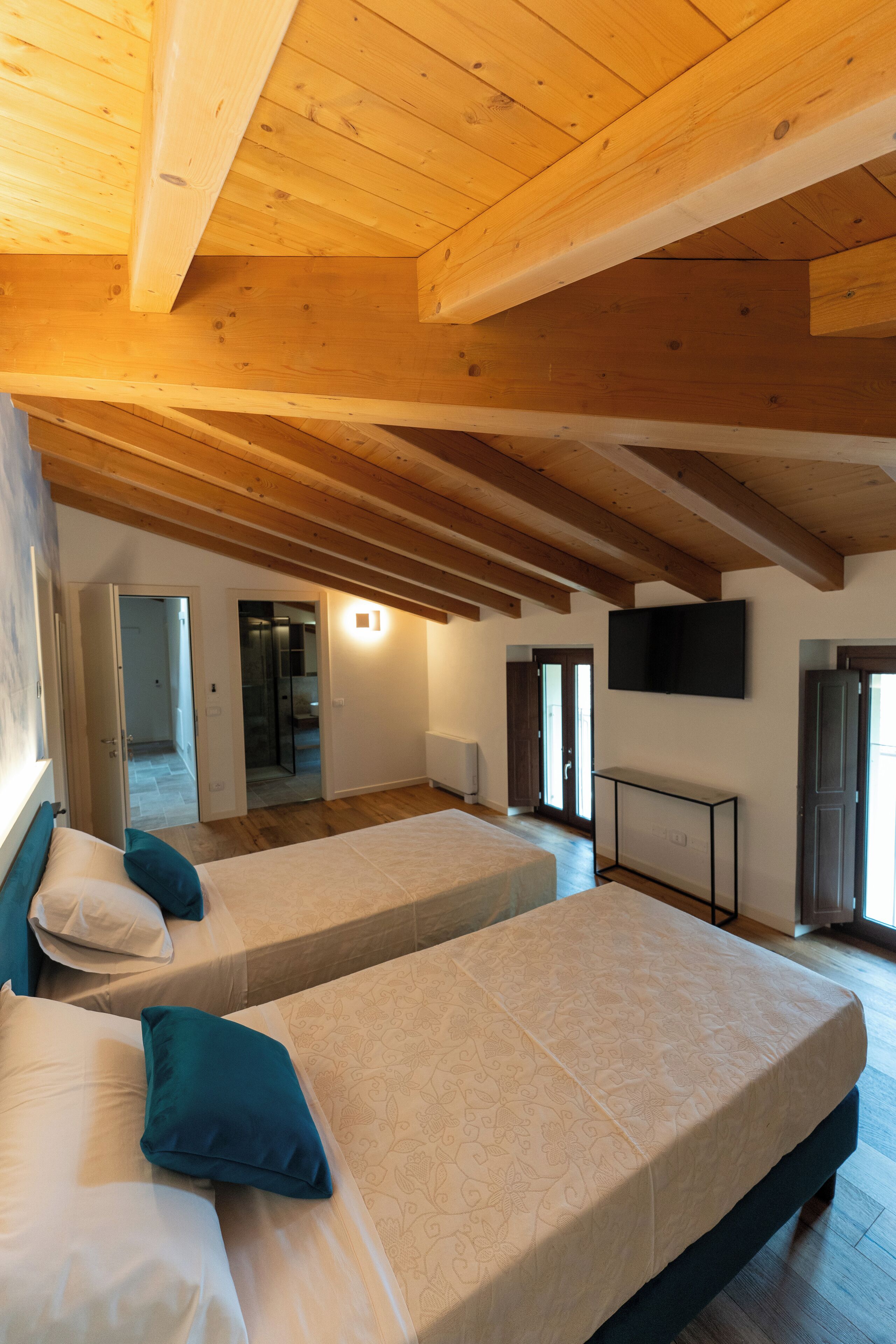 Una habitación con dos camas, vigas de madera en el techo, un televisor en la pared y una puerta que da a otra habitación.