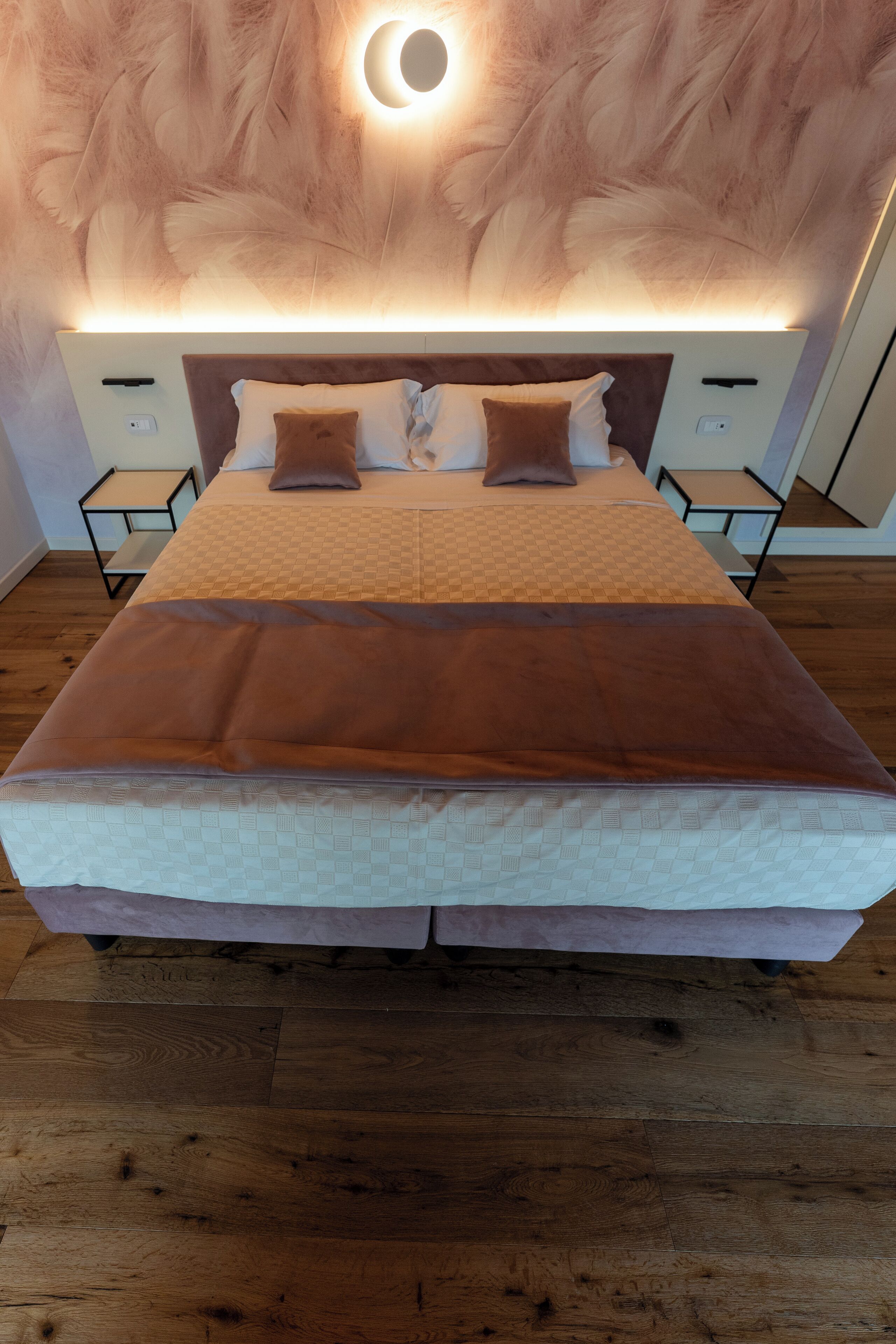 A neatly made bed with a brown blanket, a headboard with a circular light fixture, and wooden flooring.