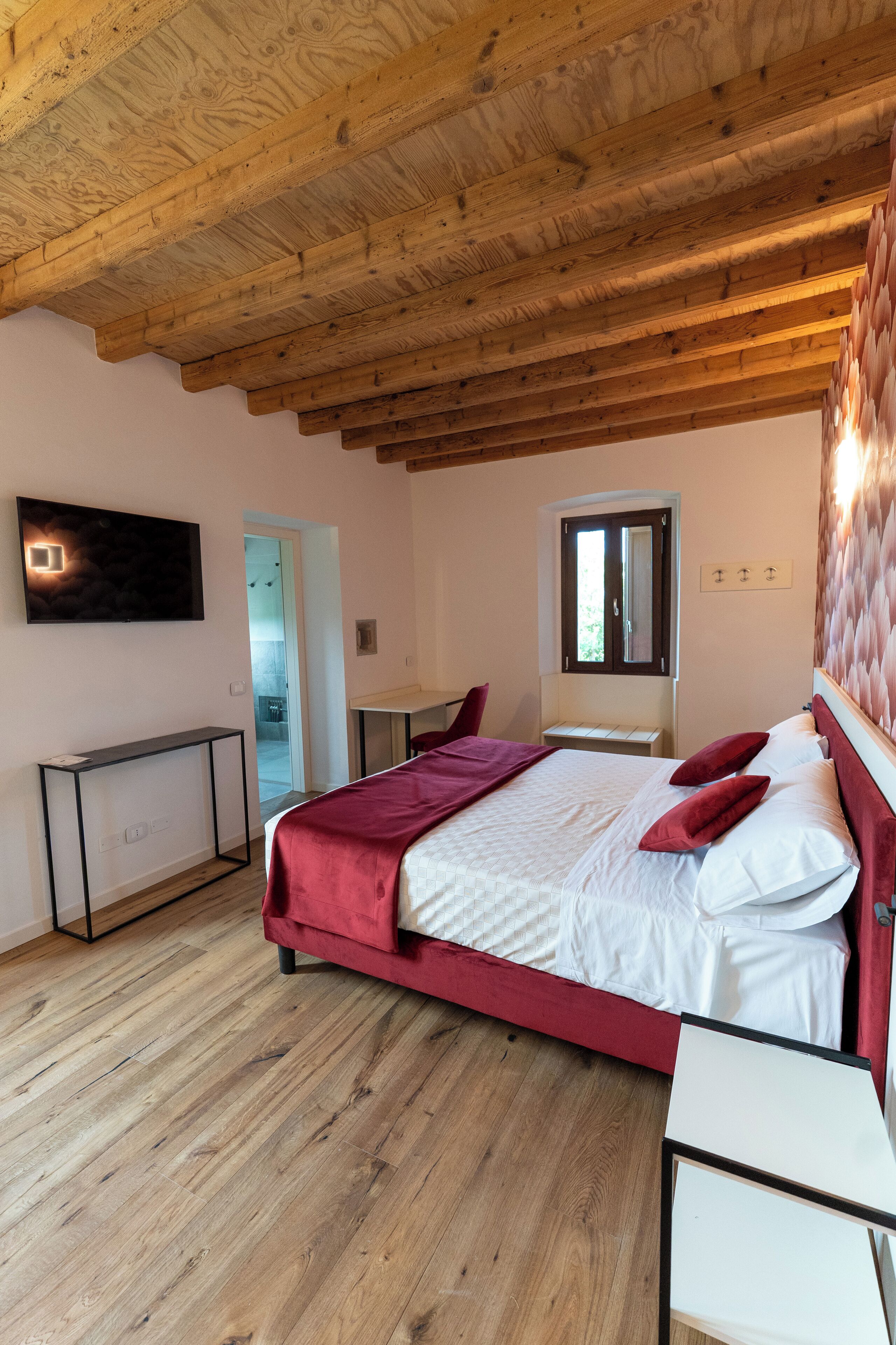 Ein Schlafzimmer mit Holzboden, einem Bett mit roter Bettwäsche, einem Nachttisch, einem an der Wand montierten Fernseher und einem Fenster mit Blick ins Freie.