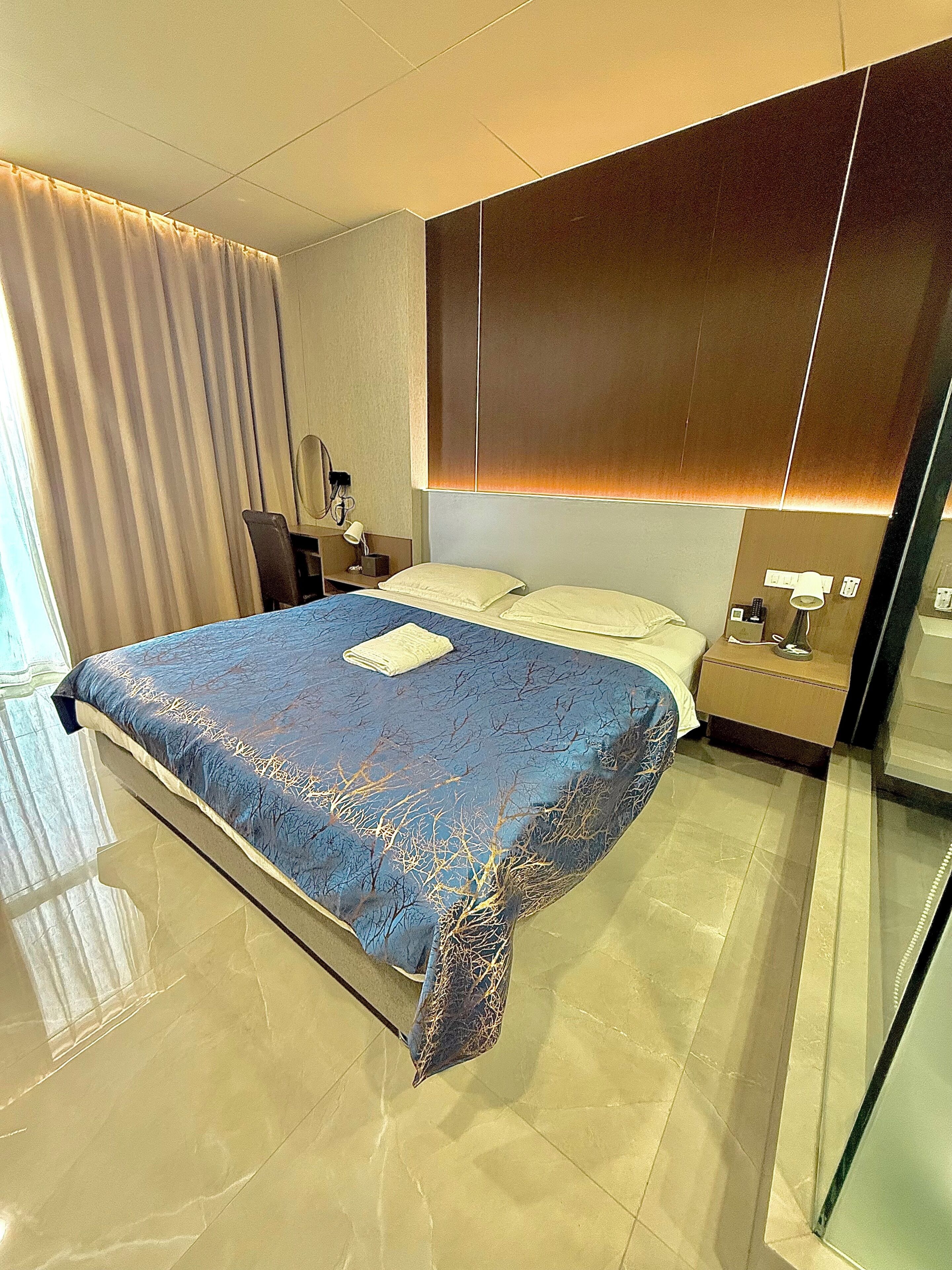 A modern hotel room with a large bed, a bedside table with a lamp, a mirror, and a window with curtains.