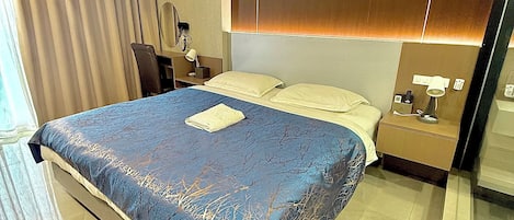 A modern hotel room with a large bed, a bedside table with a lamp, a mirror, and a window with curtains.