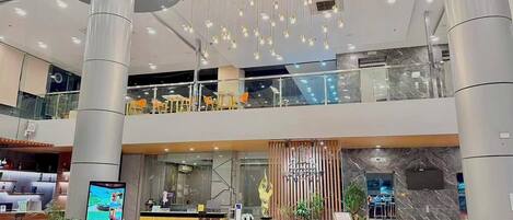 A modern hotel lobby with a reception desk, a digital information kiosk, a decorative fountain, and a glass balustrade overlooking a lower level.