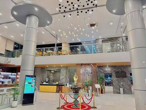 A modern hotel lobby with a reception desk, a digital information kiosk, a decorative fountain, and a glass balustrade overlooking a lower level.