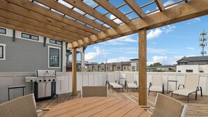 Een overdekt terras buiten met een houten tafel en rieten stoelen, een barbecue en uitzicht op omliggende huizen.