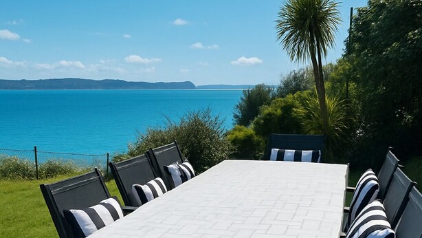 A rectangular outdoor dining table with six chairs, set on a patio with a view of the sea and hills in the background.