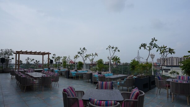 An outdoor patio with multiple seating areas, including tables and chairs, surrounded by trees and overlooking a cityscape. - Shri Rawatpura Sarkar Seva Ashram (Haridwar)