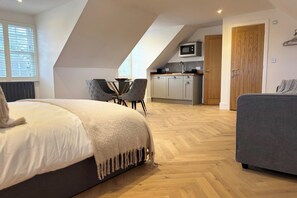 A bedroom with a bed, a grey sofa, a dining table with chairs, a kitchenette, and a door.