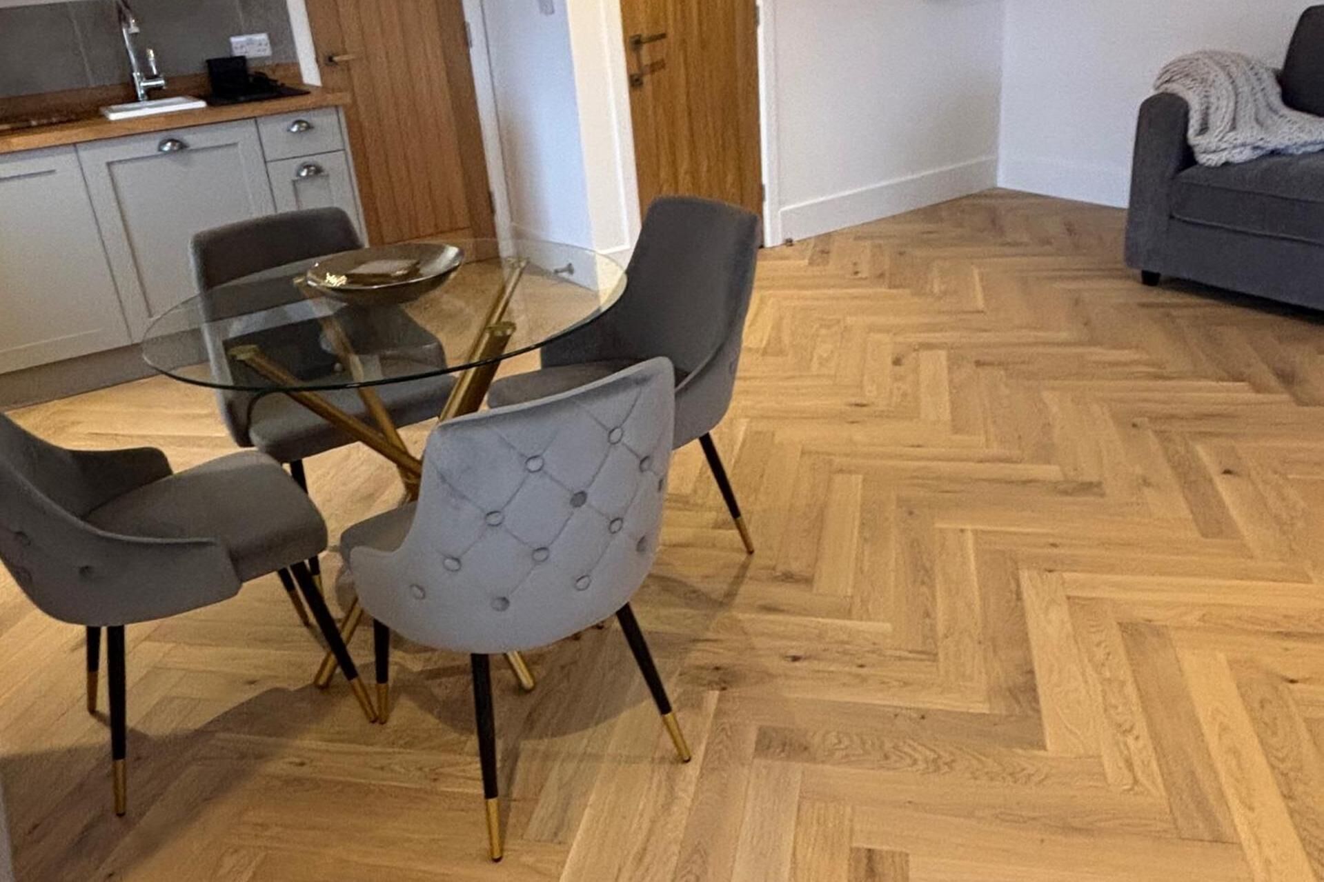 A dining area with a glass table surrounded by four grey upholstered chairs with gold legs.