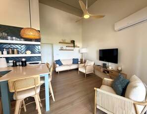 Una sala moderna con sofá, sillas, una mesa de comedor, ventilador de techo, televisor y una zona de cocina con estantes y frascos.