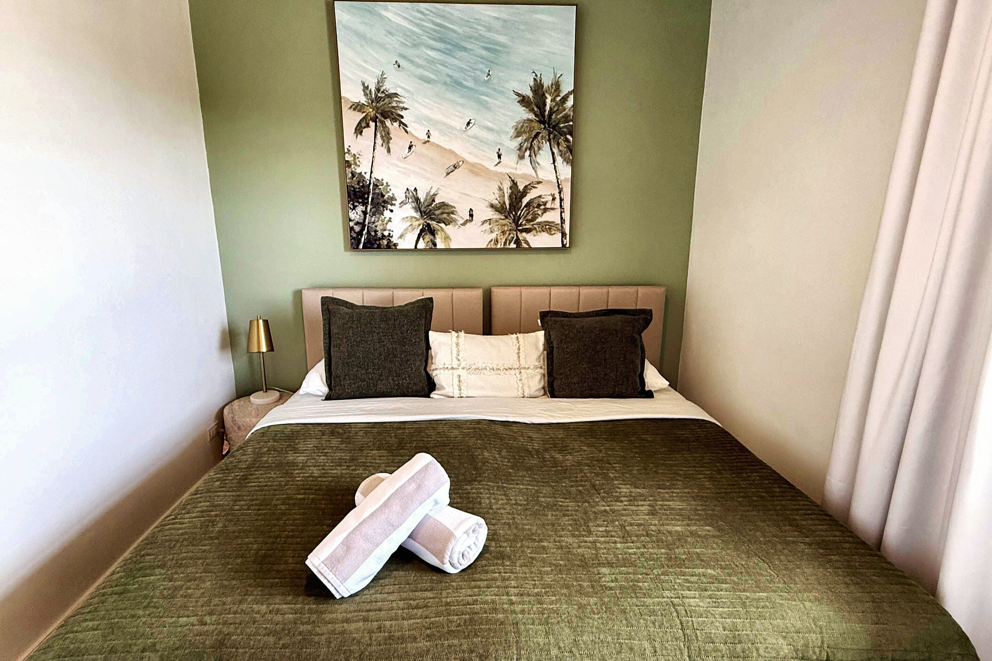 A hotel room with a bed, pillows, a painting of a beach scene, and a rolled towel.