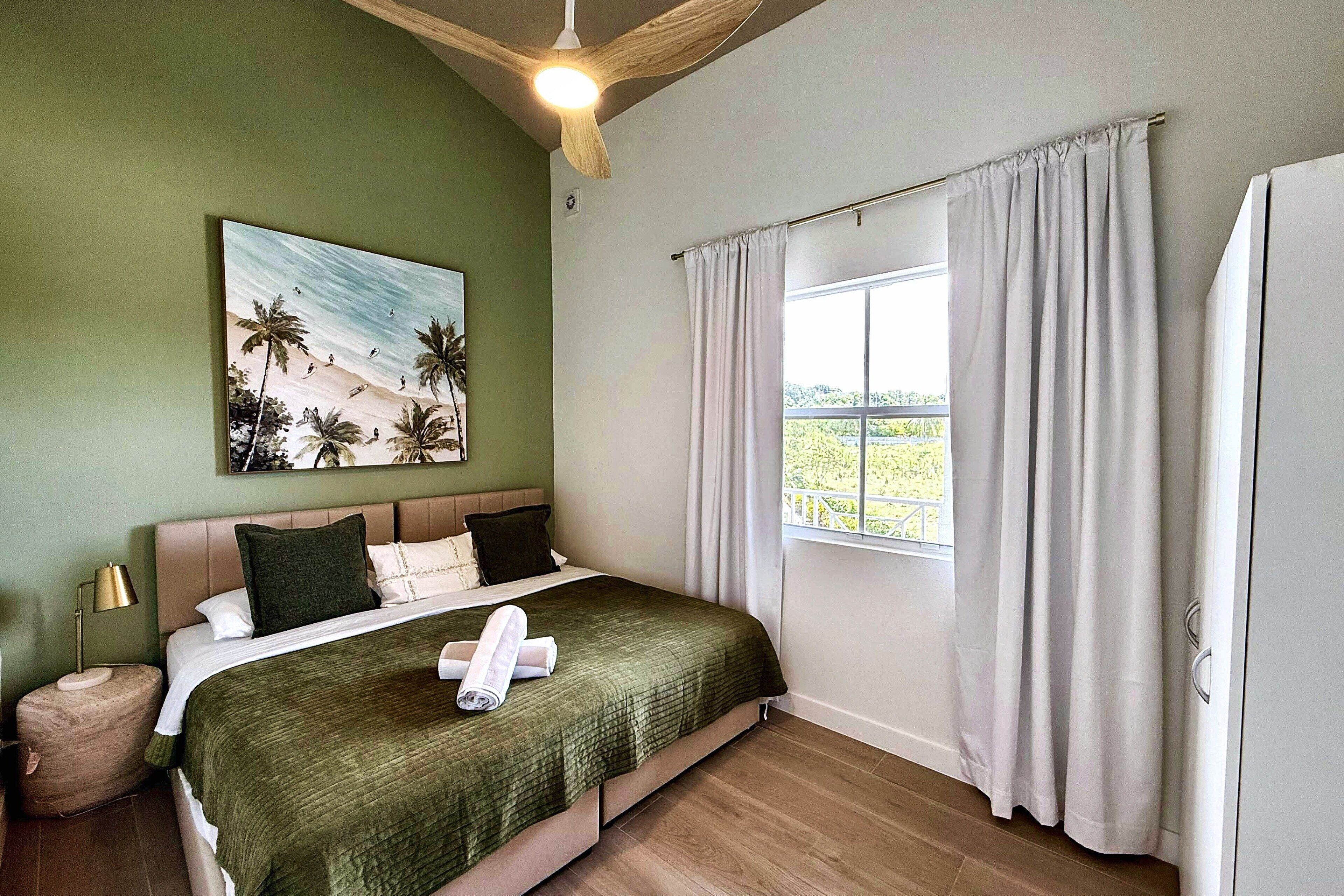 A bedroom with a bed, a painting of a beach scene, a window with curtains, and a ceiling fan.