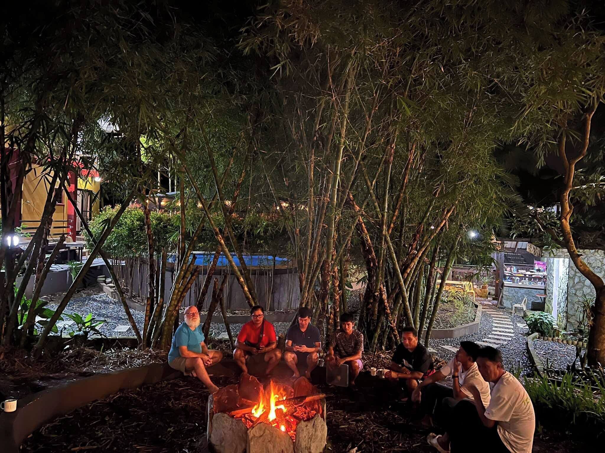 A group of people sitting around a campfire in a shaded outdoor area with trees and a building in the background.