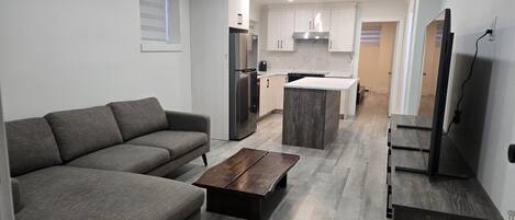 A modern kitchen with white cabinets, a grey sofa, a wooden coffee table, and a TV mounted on the wall.
