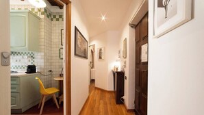 Un couloir étroit au plancher en bois, une chaise jaune et un coin cuisine avec un mur recouvert de carreaux.