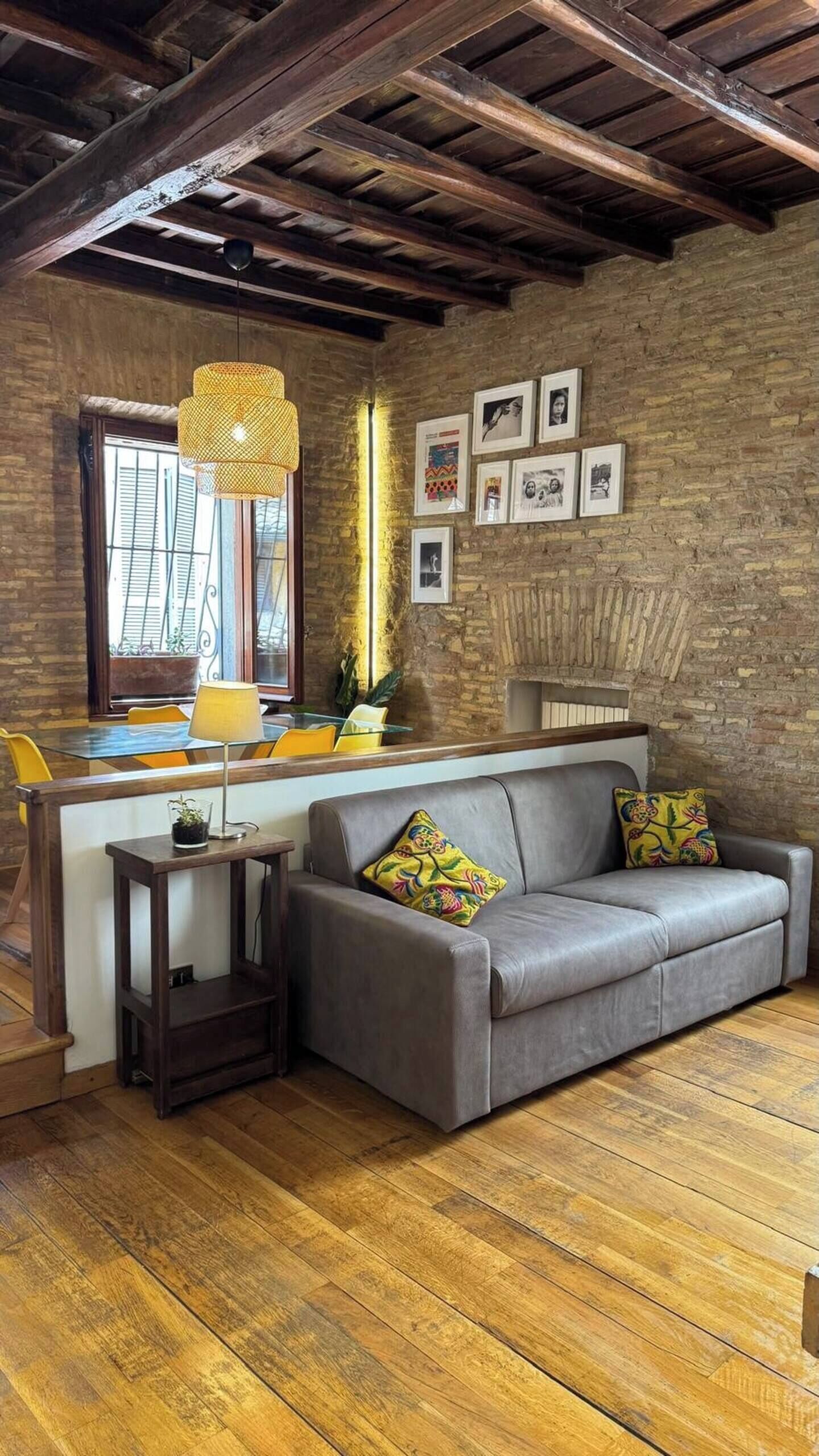 Ein gemütliches Wohnzimmer mit Ledersofa, Holzboden, einer Ziegelwand und einem Essbereich mit Glastisch und gelben Stühlen.