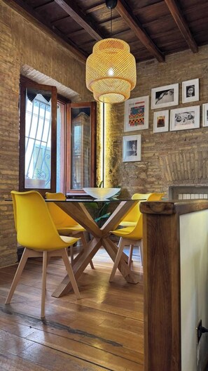 A dining area with a wooden table, yellow chairs, a woven pendant light, and a brick wall with framed pictures. - Pantheon Private Apartment (Roma)
