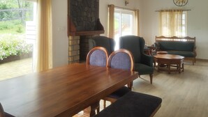 A dining area with a wooden table and chairs, a stone fireplace, and a green sofa in a well-lit room with large windows.