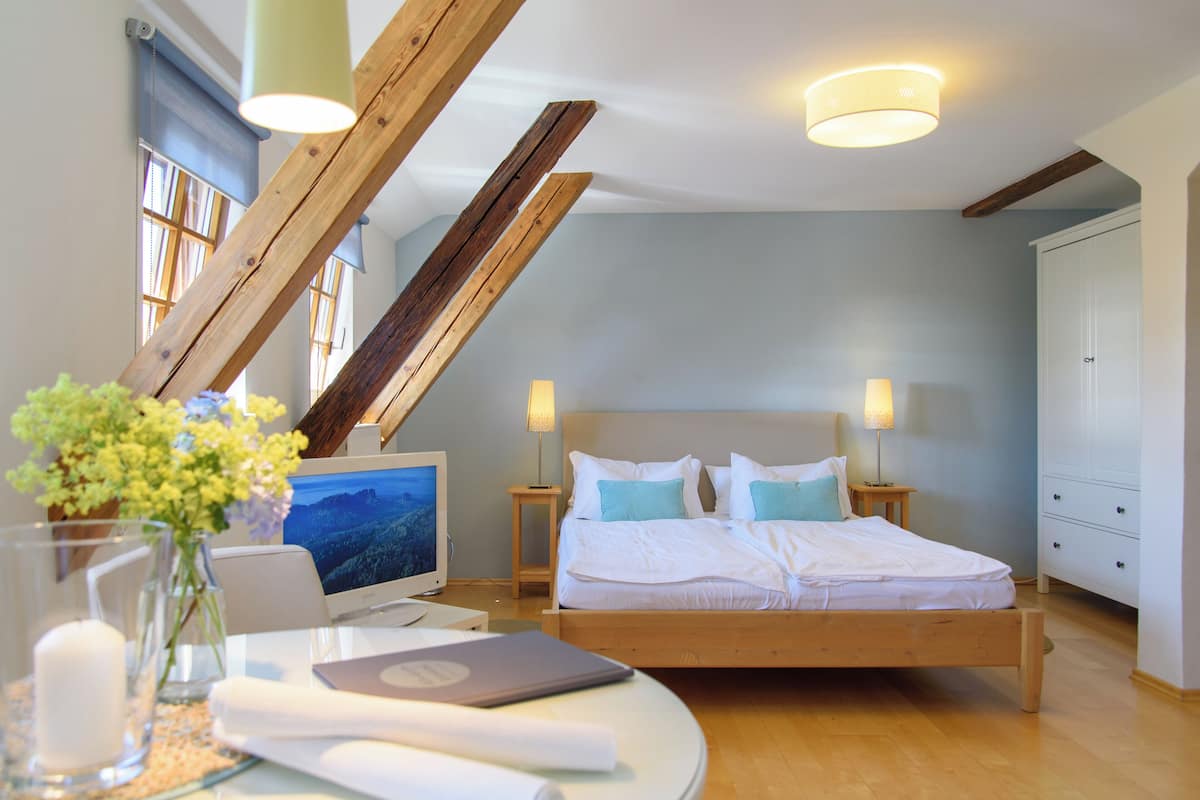 Un dormitorio con cama, lámparas de noche, un escritorio con computadora, un florero con flores y una ventana con persianas.