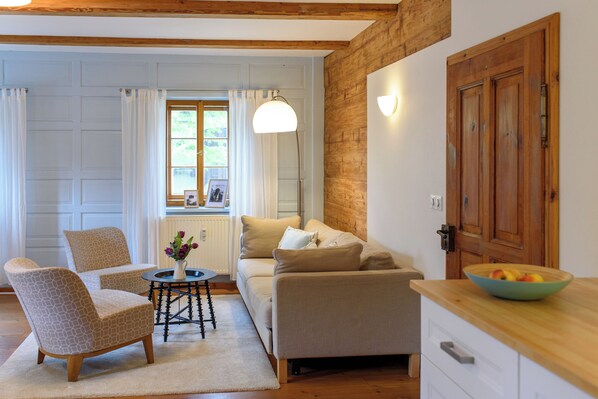 Ein gemütliches Wohnzimmer mit einer Couch, zwei Sesseln, einem kleinen Tisch mit einer Blumenvase und einer Holztür.