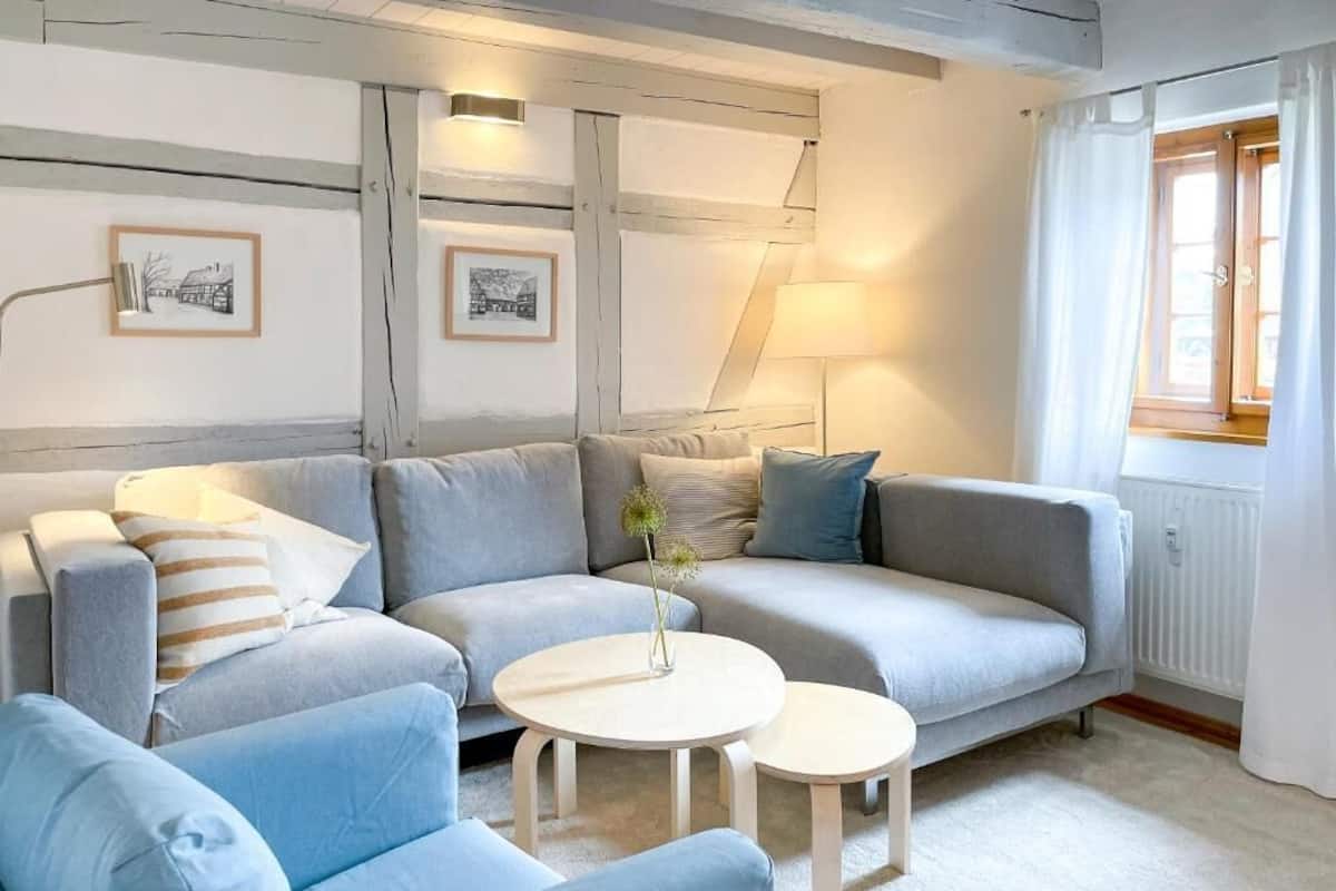 Una sala de estar moderna con un sofá gris, dos mesas redondas blancas y un ventanal con cortinas.